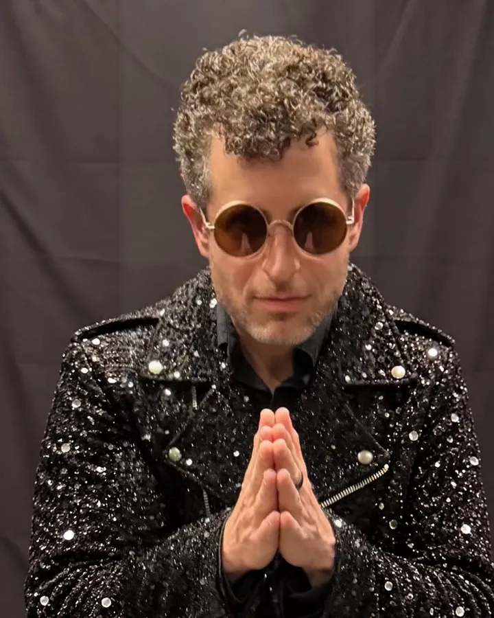 Greg Spero in round sunglasses and a sequined jacket, hands together